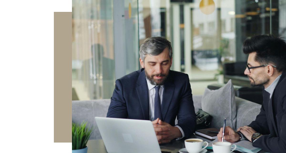 A medium close-up of two businessmen in dark suits, sitting at a table with coffee cups and a laptop. The man on the left, with a gray beard, is looking down at the open laptop screen. The man on the right, with dark hair and glasses, is facing him and writing on a paper with a pen. They appear to be in a modern, well-lit office or business lounge with glass partitions and subtle natural tones in the background.