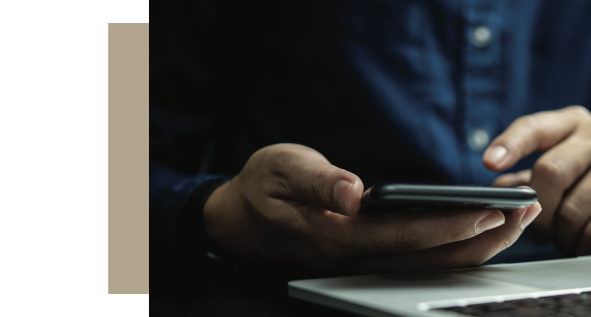 A close-up shot shows a person's hands holding a dark-colored smartphone over a laptop keyboard, likely in the process of logging in or using a two-factor authentication app.