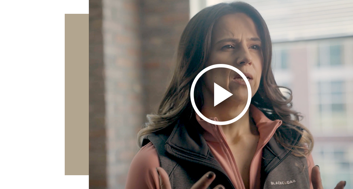 A medium close-up of a woman with long dark hair, wearing a black vest over a pink top, gesturing with her hands while speaking indoors, with a play button icon overlaid, indicating a video thumbnail.