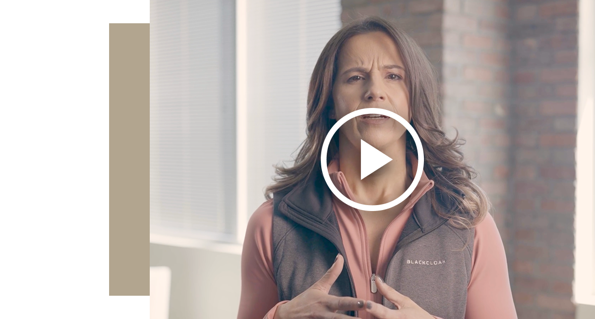 Still frame from a back-to-school cybersecurity video featuring a female speaker with dark hair in a professional, casual vest. A large white play button overlay is centered on the image.
