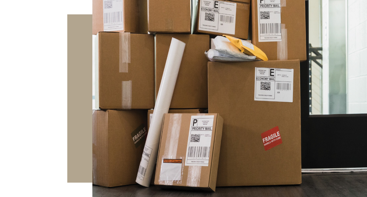 A large stack of various sizes of brown cardboard shipping boxes and an envelope, some labeled with Priority Mail and Economy Mail stickers, are piled up near an entryway. One box has a red "FRAGILE" sticker.