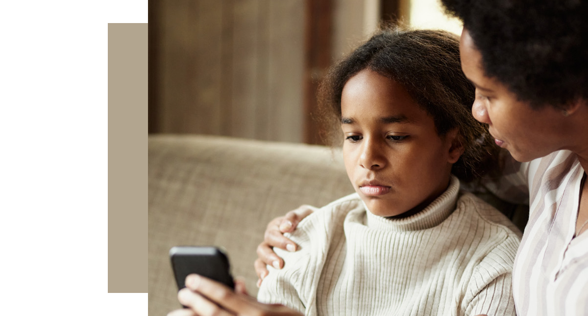 Close-up of a young girl and an adult woman, likely a mother, sitting on a couch and looking intently at a smartphone. The image conveys a serious discussion about using the internet safely.