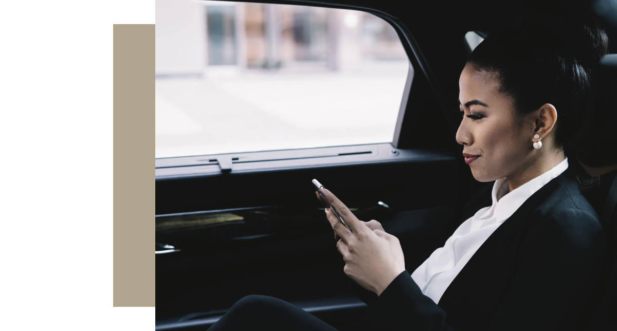 A professional woman in a business suit sitting in the back seat of a car, looking down and interacting with her cell phone.