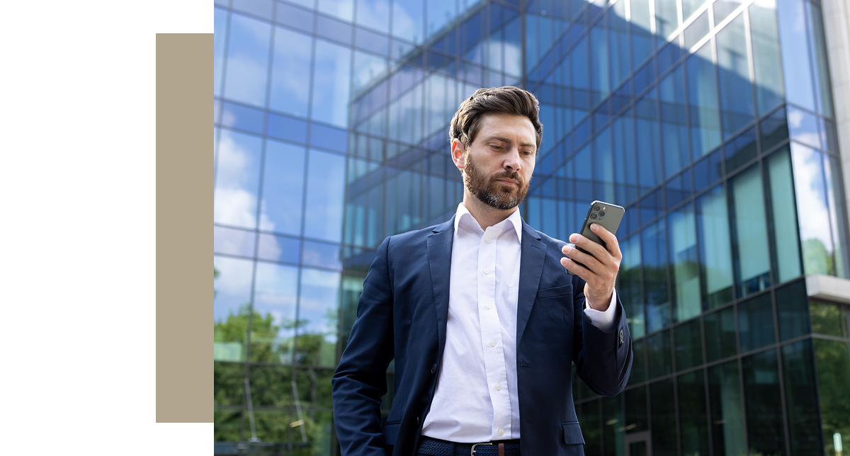A man in a suit stands outside a modern glass office building, looking intently at his smartphone. He has a concerned or focused expression.