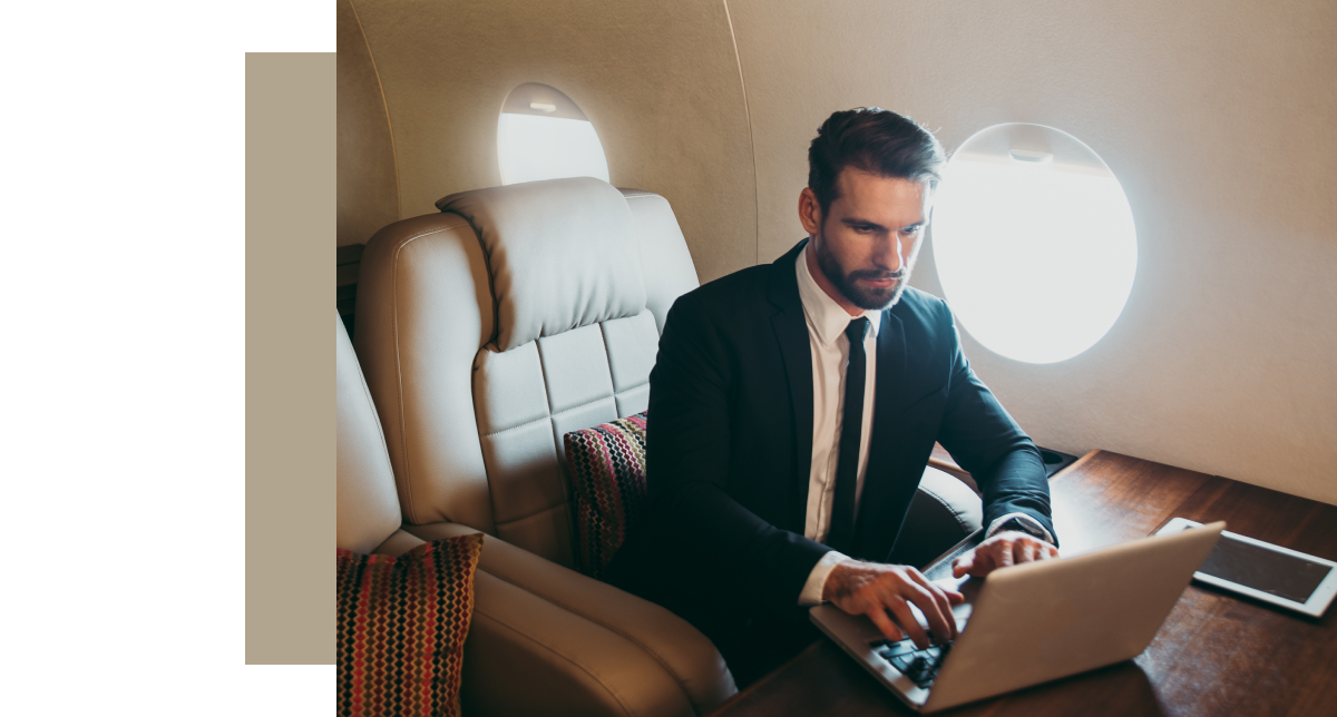 A man in a suit is sitting in a luxurious private jet cabin, focusing on working on a laptop on a wooden table.