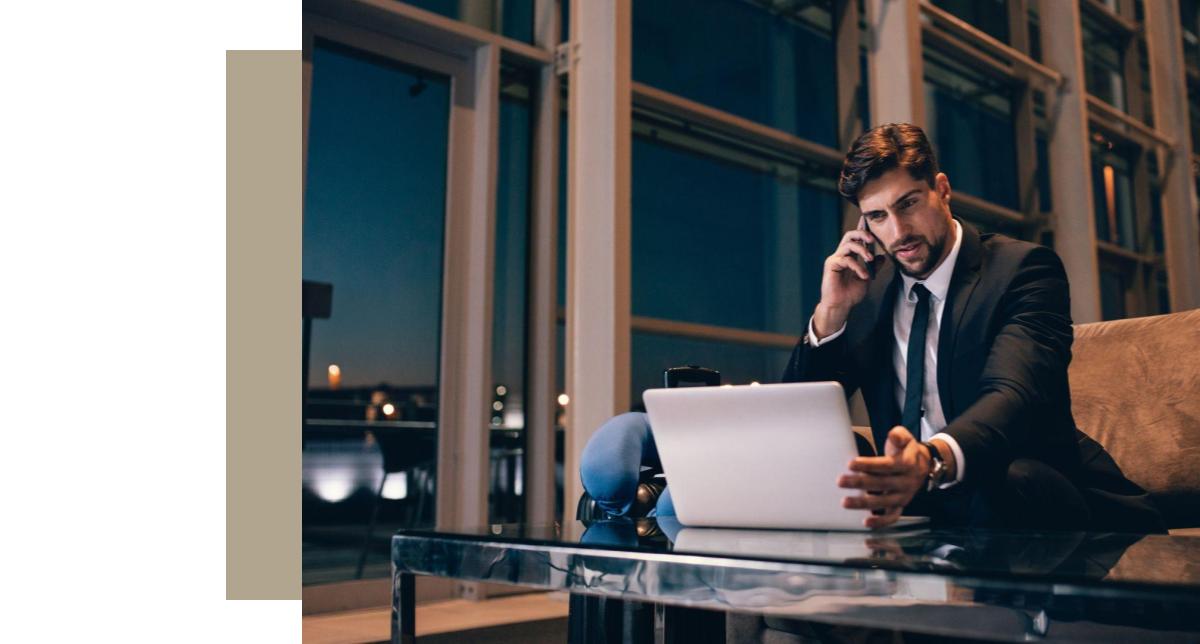 A man in a dark suit and tie is seated at a low glass table, looking intently at a laptop screen while holding a phone to his ear. He is in an upscale, modern interior space with large windows overlooking a city view at night. A beige and white color block graphic is on the left side of the image.