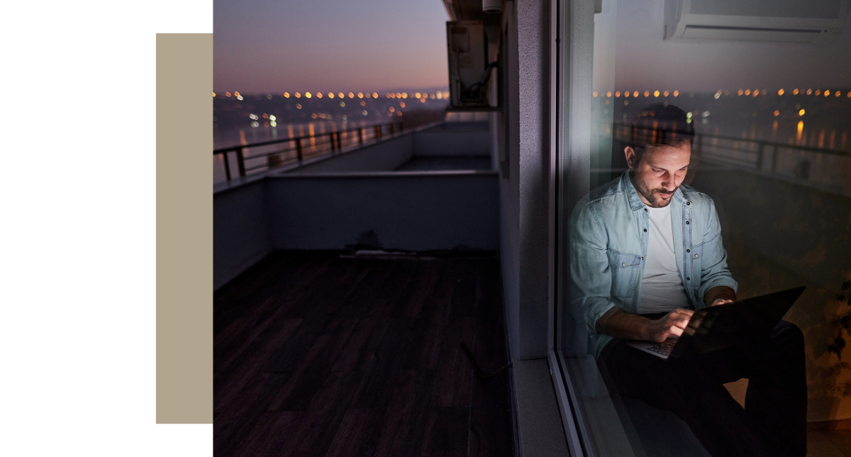 Man working on laptop at night by a glass door overlooking a city skyline
